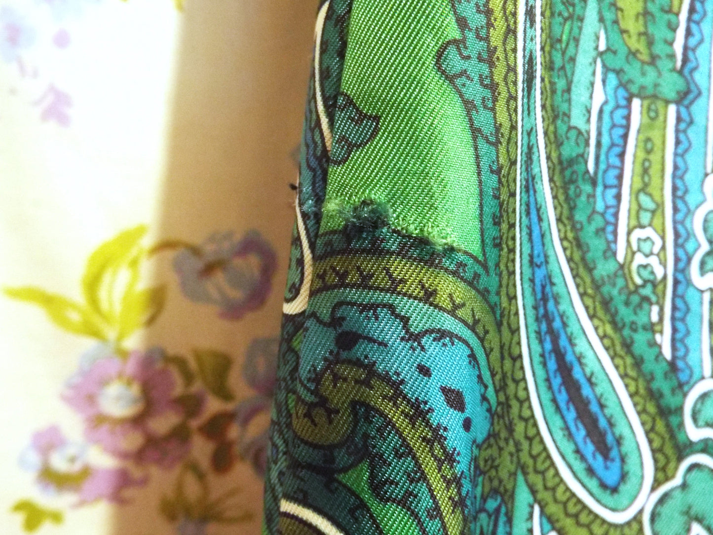 Close-up of pull in fabric on Vintage Handmade Paisley Dress (S-M) in a green and black paisley print, against patterned fabric with blue and purple roses.