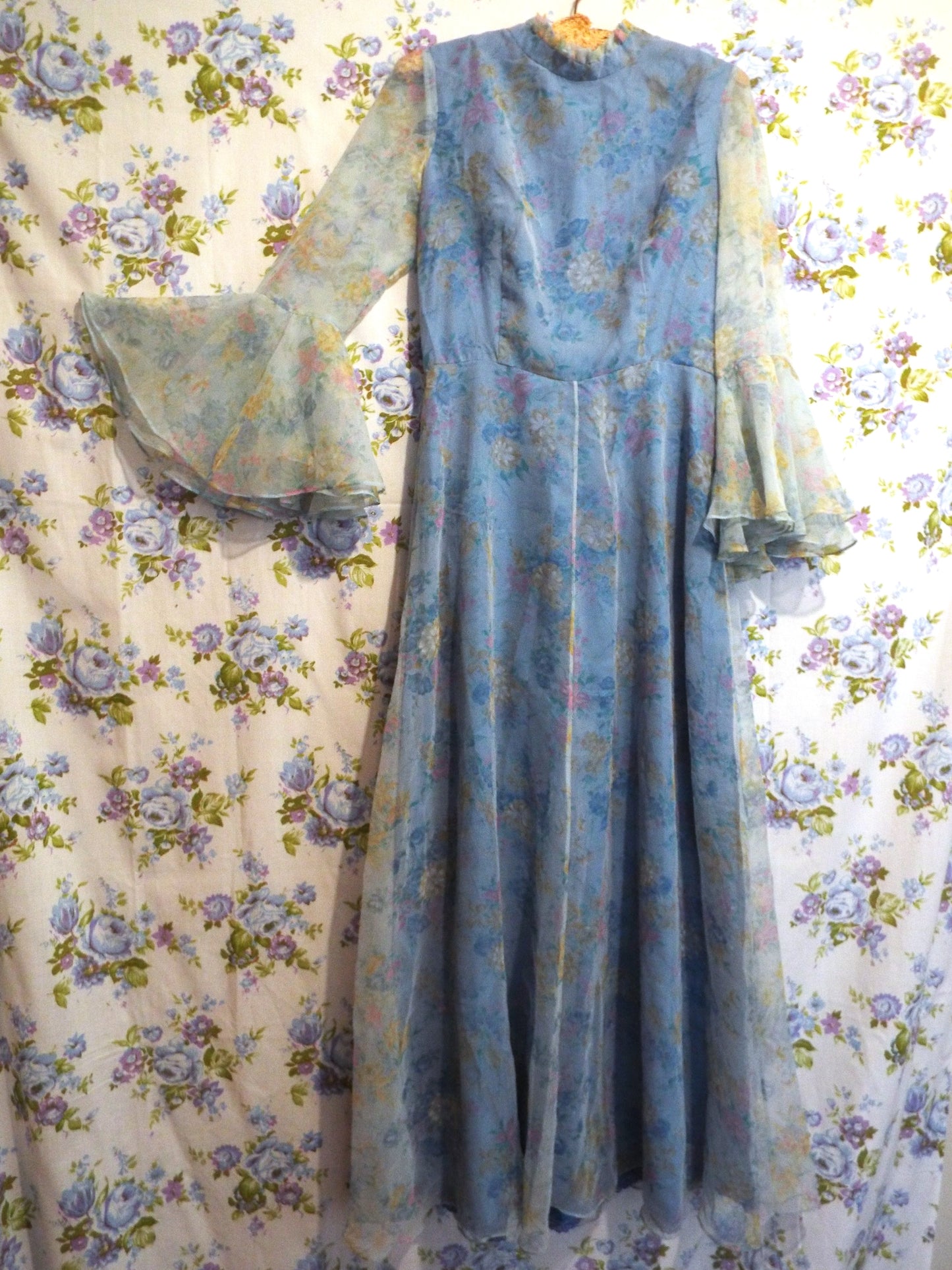 1970s Maxi Dress (S-M) with light blue floral patterned fabric and puffy sleeves, against patterned fabric with blue and purple roses.