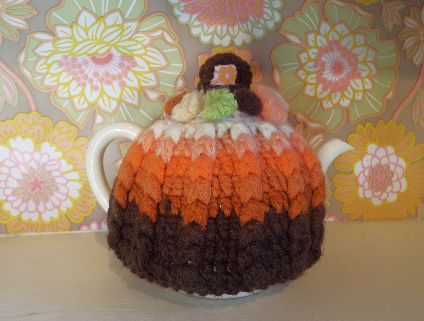 Small Vintage Tea Cosy with brown and orange ombre design against warm floral backdrop.