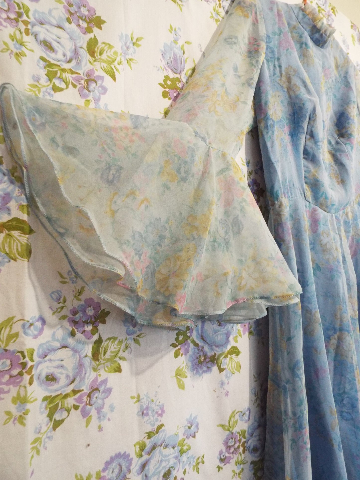 1970s Maxi Dress (S-M) with light blue floral patterned fabric and puffy sleeves, against patterned fabric with blue and purple roses.